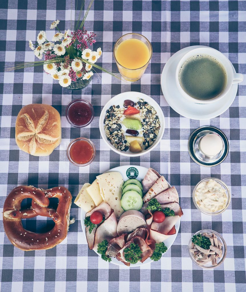 Auf grau/wei karierter Tischdecke: Teller mit Wurst, Käse, Gemüsedeko, Breze, Semmel, 2 Schalen mit Marmelade, Müslischale mit Obst, Ei im eierbecher, Tasse Kaffee, Saftglas, Vase mit Blumen