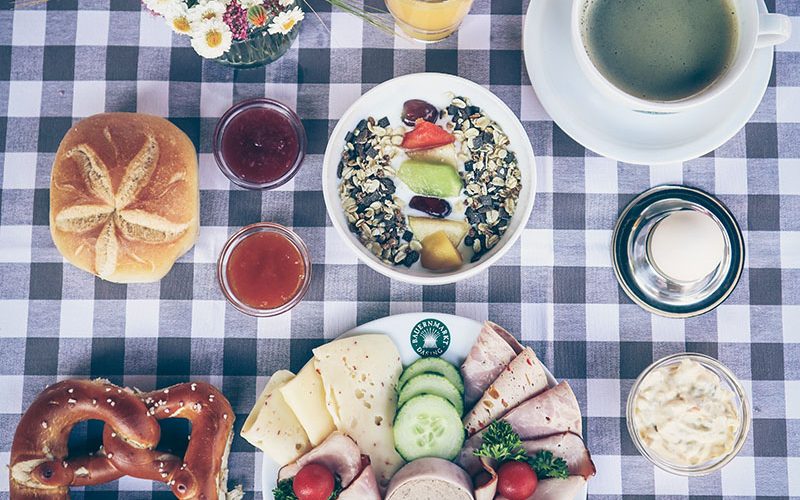 Frühstücksteller mit Wurst, Käse, Gurke, Müslischale mit buntem Obst, Schälchen mit salaten, Kaffee, Saftglas, Breze, Semmel, Blumenvase auf grau karierter Tischdecke