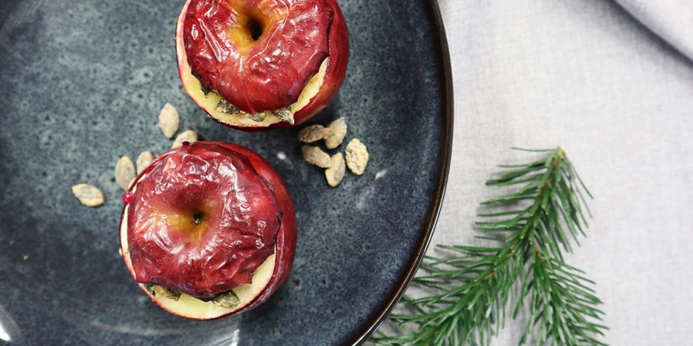 zwei Bratäpfel auf dunklem Teller auf grauer Decke daneben ein Tannenzweig